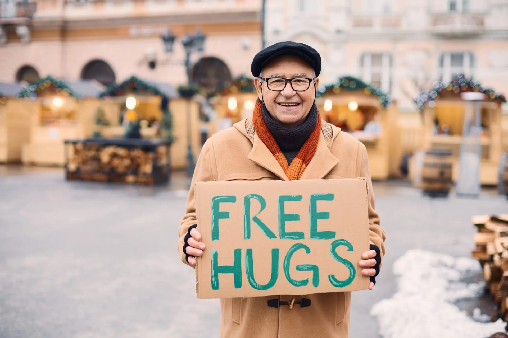 Smiling older man in winter clothes and scarf stands in a city square smiling a holding a "Free Hugs" sign.